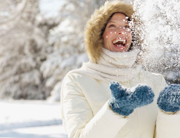 Frau in Winterjacke und Handschuhen wirft lachend Schnee in die Luft.