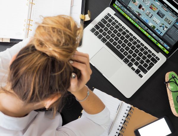 Frau arbeitet am Laptop, legt Hände an den Kopf, neben ihr liegen Ordner und Notizen
