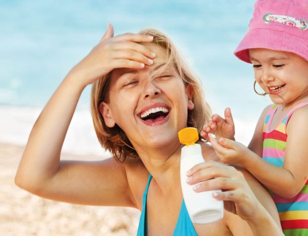 Mutter und Kind mit Sonnenhut lachen am Strand als Symbol für Sonnenschutz.