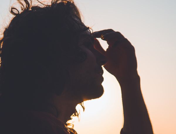 Frau hält sich den Kopf mit beiden Händen, angestrahlte Silhouette bei Sonnenuntergang als Darstellung von Kopfschmerzen und Migräne