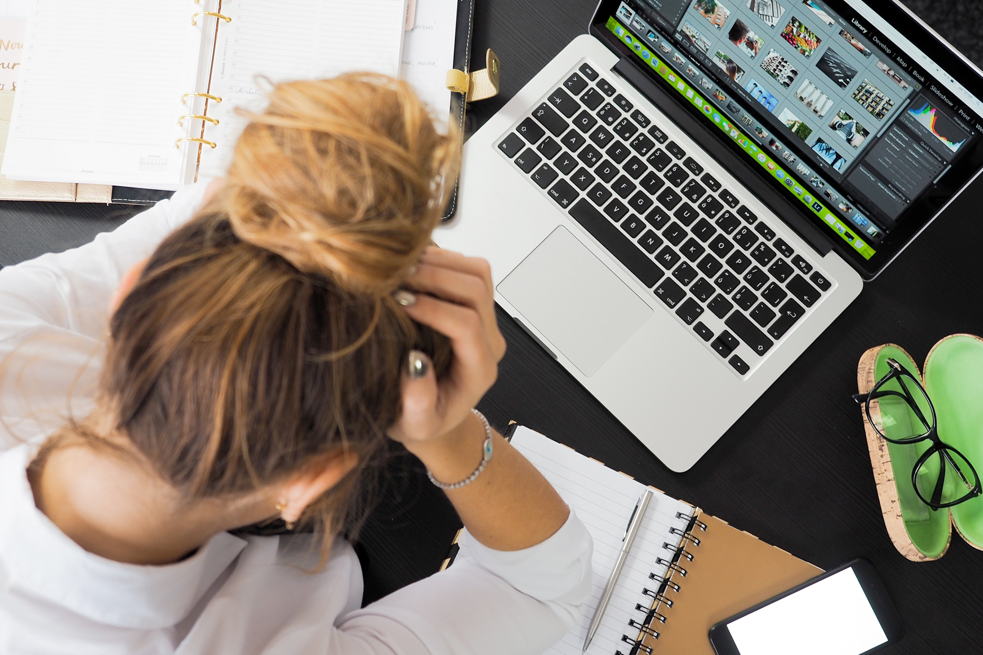 Frau arbeitet am Laptop, legt Hände an den Kopf, neben ihr liegen Ordner und Notizen