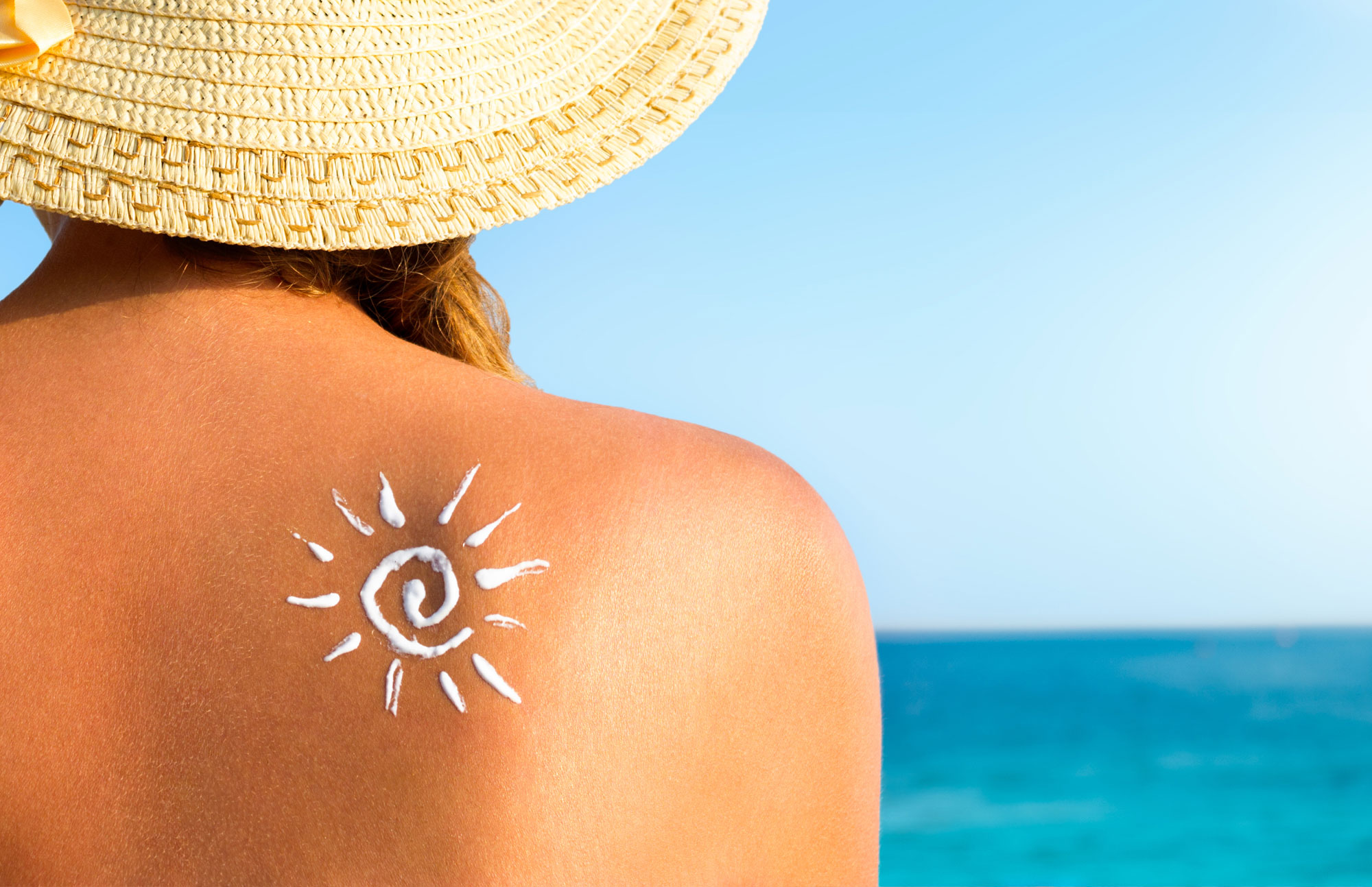 Frau mit Sonnenhut sitzt am Strand, auf der Schulter ist ein Sonne-Symbol aus Sonnencreme aufgetragen.