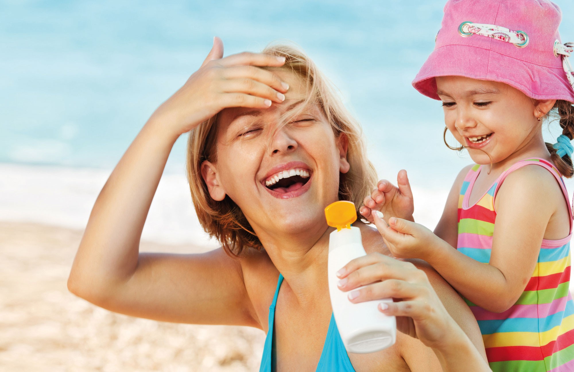 Mutter und Kind mit Sonnenhut lachen am Strand als Symbol für Sonnenschutz.
