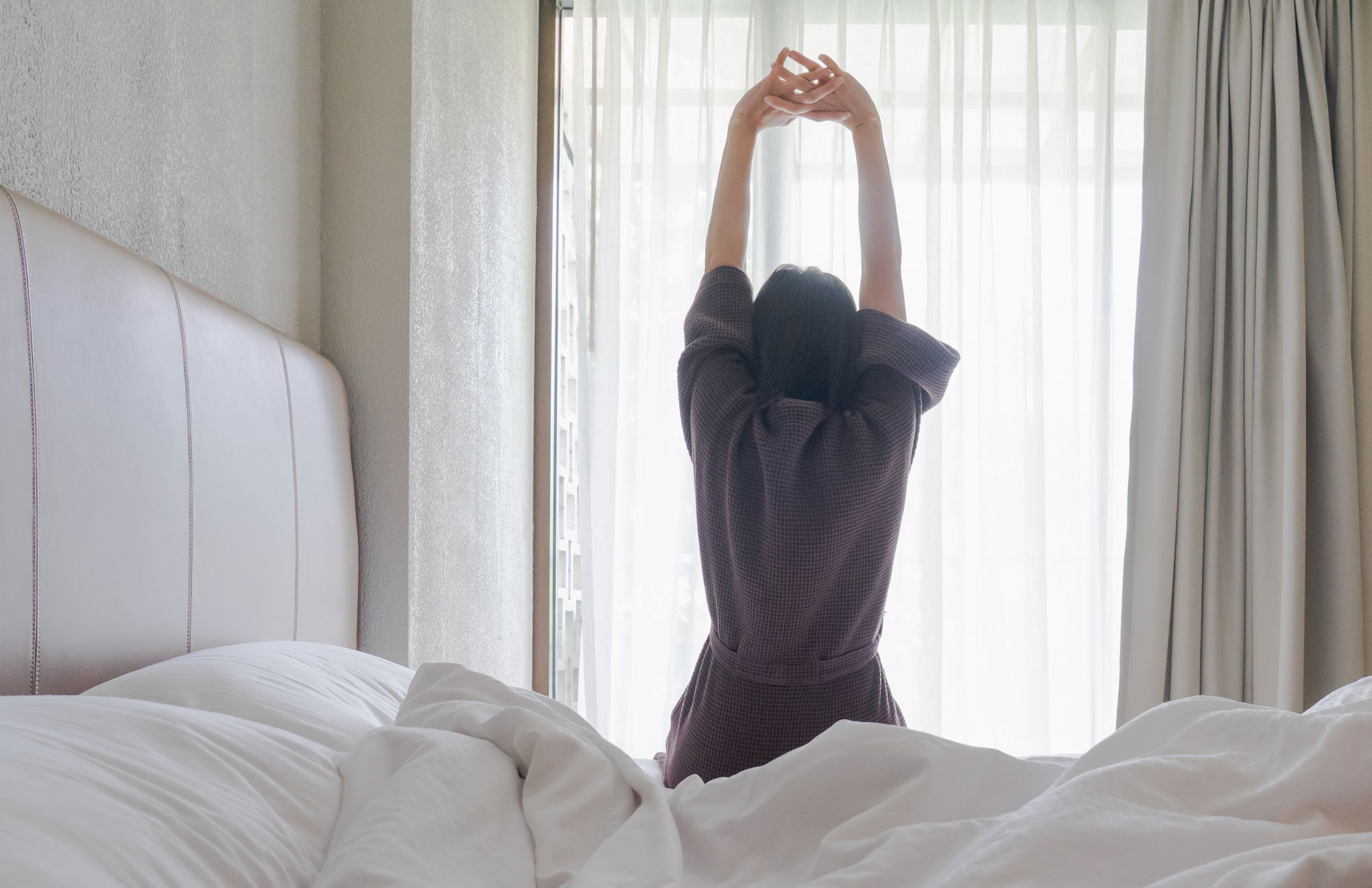 Frau sitzt im Bett und streckt beim Aufwachen die Arme nach oben.